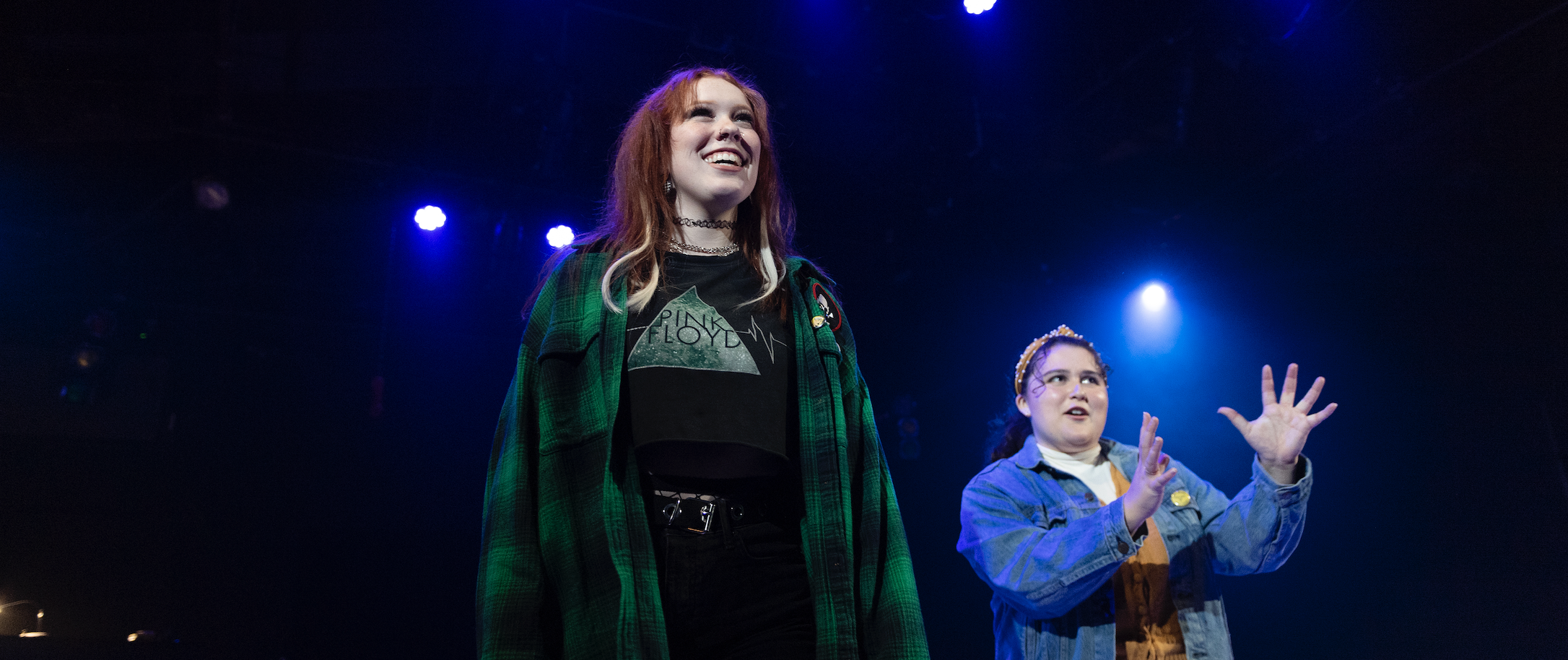 A young white woman with red hair and a green plaid shirt looks up while a Latinx woman with curly brown hair and a blue jean jacket gestures up and out