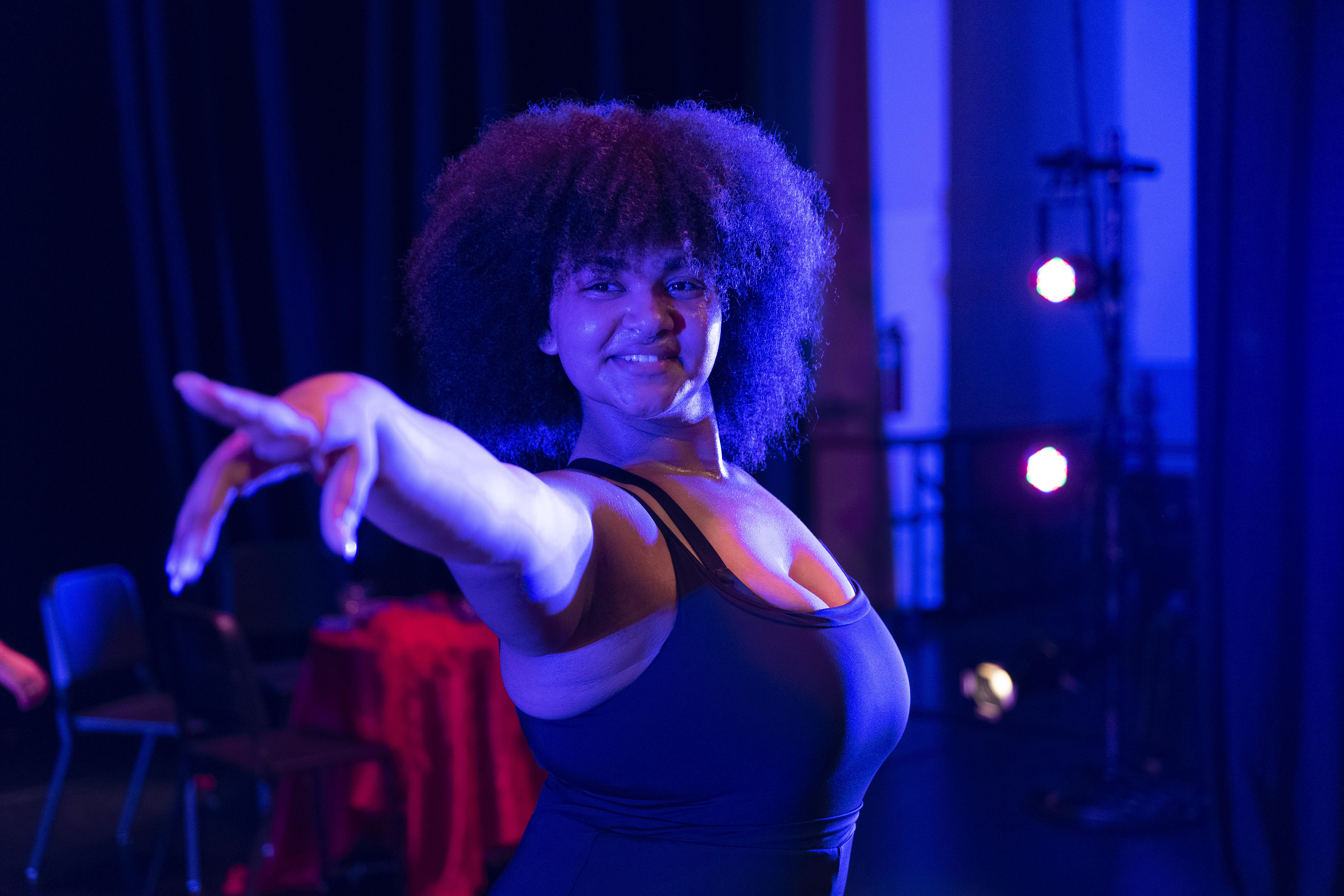 A young Black woman wears a black leotard flexes her fingers towards the camera