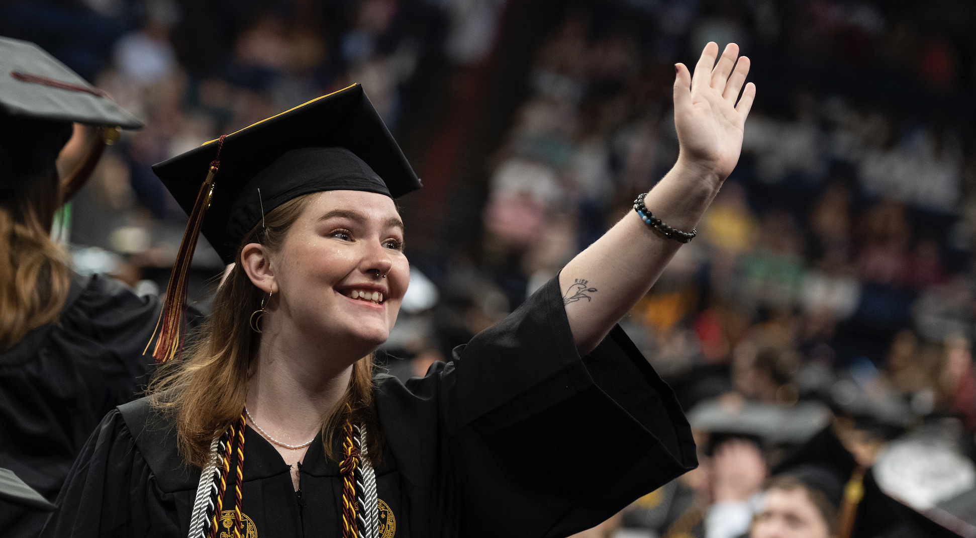 A student waves to someone out view. They are wearing a black graduation cap and gown.